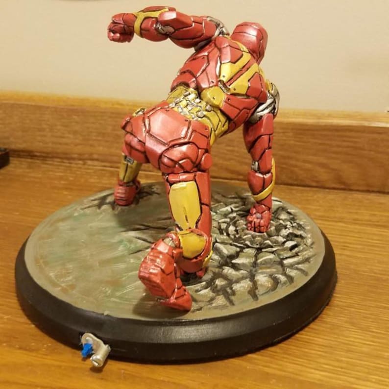 May include: A red and gold Iron Man figurine in a dynamic pose, set on a round, textured base. The figurine's armor features detailed paneling and a metallic sheen. A small, detached piece lies near the base.