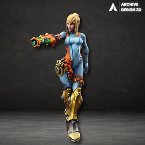 May include: A 3D-printed figurine of a female character in a futuristic suit. The figure is predominantly light blue with orange and gold accents. The character is holding a large weapon. The logo "ARCHIVE DESIGN 3D" is in the top right corner.