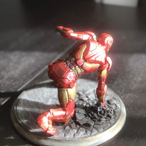 May include: A red and gold Iron Man figurine in a kneeling pose. The figurine is on a circular base with a textured surface. The figure's armor has intricate details and a metallic sheen, with gold accents.