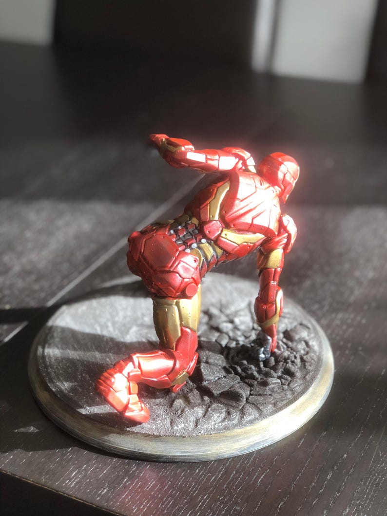May include: A red and gold Iron Man figurine in a kneeling pose. The figurine is on a circular base with a textured surface. The figure's armor has intricate details and a metallic sheen, with gold accents.