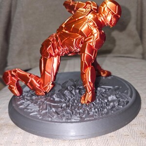 May include: A 3D-printed figurine of a superhero in a crouching pose. The figure is a metallic orange color with detailed armor plating. It is mounted on a gray, textured base with a circular border, suggesting a dynamic action scene.