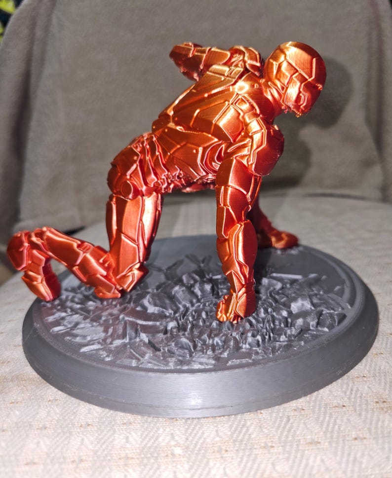 May include: A 3D-printed figurine of a superhero in a crouching pose. The figure is a metallic orange color with detailed armor plating. It is mounted on a gray, textured base with a circular border, suggesting a dynamic action scene.