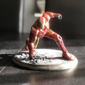 May include: A red and gold Iron Man action figure in a dynamic pose, set on a gray, textured base. The figure is detailed with metallic accents and stands approximately 6 inches tall.