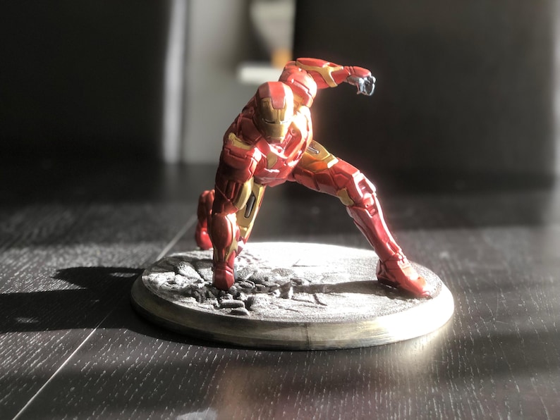 May include: A red and gold Iron Man action figure in a dynamic pose, set on a gray, textured base. The figure is detailed with metallic accents and stands approximately 6 inches tall.