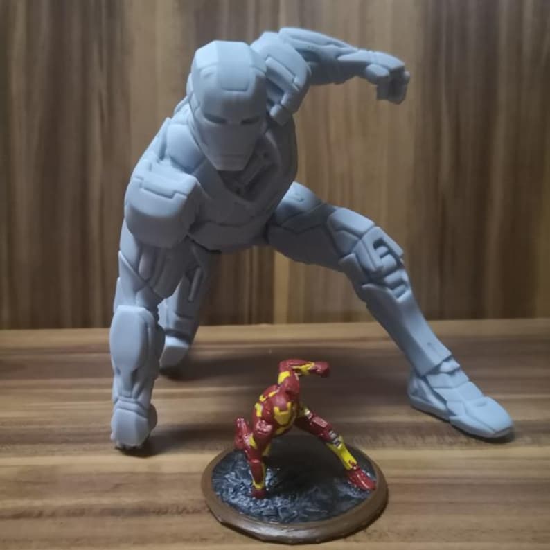 May include: A large, gray Iron Man figurine in a dynamic pose, with one arm extended and the other bent. A smaller, red and gold Iron Man figure is positioned in front of it on a circular base. The figures are on a wooden surface.