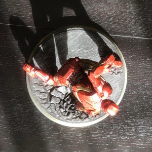 May include: A red and gold Iron Man action figure in a dynamic pose, set on a circular base with a textured, rocky surface. The figure is highly detailed, with visible armor plating and a metallic sheen. The base is approximately 4 inches in diameter.