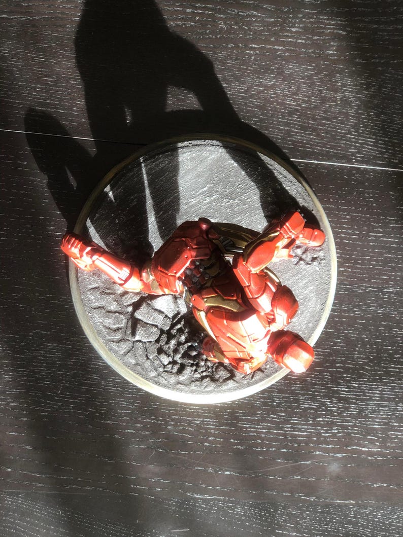 May include: A red and gold Iron Man action figure in a dynamic pose, set on a circular base with a textured, rocky surface. The figure is highly detailed, with visible armor plating and a metallic sheen. The base is approximately 4 inches in diameter.