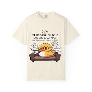 May include: Cream-colored t-shirt featuring a cartoon rubber duck wearing glasses, sitting on a brown couch. The text reads "RUBBER DUCK • DEBUGGING • My Therapist Has a Bill" with coding symbols in speech bubbles. A fun, geeky design.
