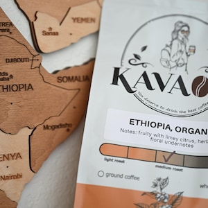 Könnte beinhalten: Eine Packung KAVAOLI äthiopischen Bio-Kaffees mit einer Afrika-Karte im Hintergrund. Die Kaffeepackung hat ein weißes und orangefarbenes Design mit dem Text "ETHIOPIA, ORGANIC" und einer Beschreibung des Geschmacksprofils des Kaffees.