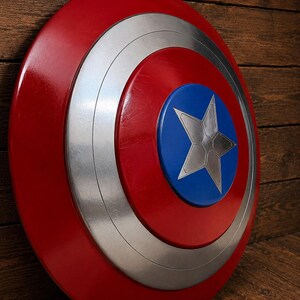 May include: A red, silver, and blue shield with a star emblem. The shield has a circular design with concentric rings of red, silver, and blue. A silver star is centered on the blue circle. The shield is displayed against a wooden background.