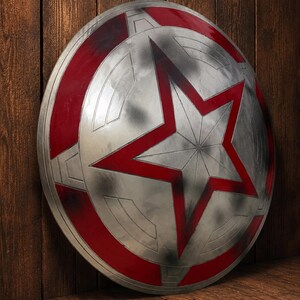 May include: A silver and red shield with a large red star in the center. The shield has a circular design with red accents around the edges. The shield is leaning against a wooden surface.