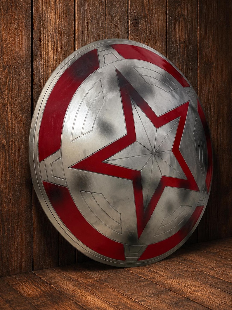 May include: A silver and red shield with a large red star in the center. The shield has a circular design with red accents around the edges. The shield is leaning against a wooden surface.