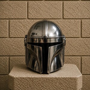 K&ouml;nnte beinhalten: Ein polierter, metallischer Mandalorianer-Helm mit einem markanten T-f&ouml;rmigen Visier. Der Helm wird auf einem strukturierten, hellbraunen Steinblock vor einer Backsteinmauer pr&auml;sentiert. Die Oberfl&auml;che des Helms reflektiert das Licht.