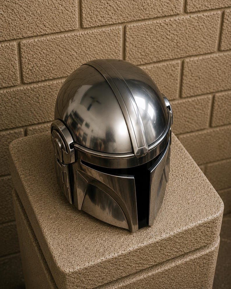 K&ouml;nnte beinhalten: Ein gl&auml;nzender, silberfarbener Helm mit einem markanten Visier und einer abgerundeten Oberseite. Der Helm hat detaillierte Rippen und eine glatte, reflektierende Oberfl&auml;che. Er liegt auf einer strukturierten, beigefarbenen Oberfl&auml;che.