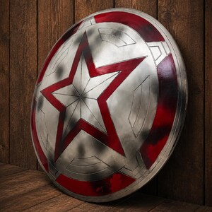 May include: A silver shield with a large red star in the center and red accents around the edge. The shield is leaning against a wooden wall and floor, creating a rustic backdrop.
