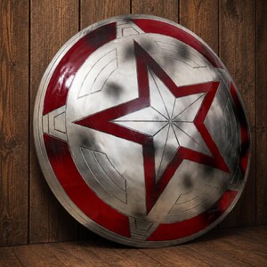 May include: A silver and red shield with a large red star in the center. The shield has a circular design with red accents and a weathered appearance. The shield is leaning against a wooden surface.