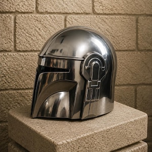 K&ouml;nnte beinhalten: Ein gl&auml;nzender, silberner Helm mit Visier und markanten Seitendetails, der auf einem strukturierten, hellbraunen Block ruht. Das Design des Helms weist eine glatte, metallische Oberfl&auml;che auf, die das Licht reflektiert. Der Helm ist ein Requisit oder Sammlerst&uuml;ck.