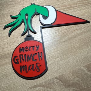 May include: A 3D printed wall decoration featuring a green hand wearing a red and white Santa hat, holding a red circular ornament. The ornament has the black embossed text merry GRINCH mas. The design is outlined in black plastic throughout.