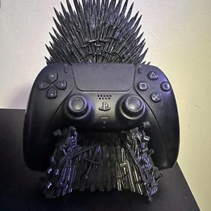 May include: A black PlayStation 5 controller on a black throne-shaped stand. The stand is a detailed replica of a throne with sharp spikes. The controller has the standard button layout and is a sleek, modern design.