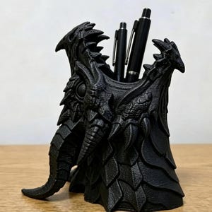 May include: A matte black desk organiser shaped like a dragon head with intricate scale textures and sharp horn details. The hollow top section holds two black pens. The structure features a wide base and a textured surface with layered scales.