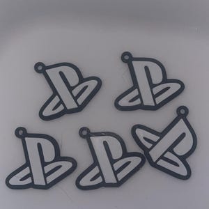 May include: Five white and black PlayStation logo charms. Each charm features the iconic PlayStation symbol with a small hole at the top for attaching to a keychain or other craft project. The charms are set against a white background.