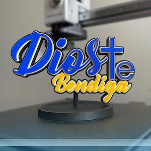 Puede incluir: Un letrero impreso en 3D sobre una base circular negra que presenta el texto azul "Dios" con una cruz reemplazando la letra "t" y el texto amarillo "Bendiga" debajo. Las letras tienen contornos superpuestos en negro y blanco.