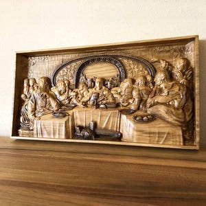 May include: A wooden relief carving depicting the Last Supper. The artwork is framed and features detailed figures seated around a table. The carving is in shades of brown and tan, showcasing the wood grain.
