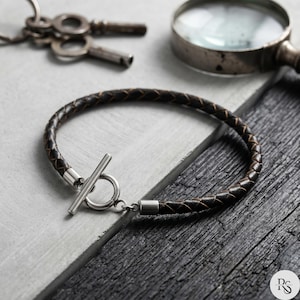 May include: A dark brown braided leather bracelet with a silver clasp. The bracelet is displayed on a gray and black wooden surface, with keys and a magnifying glass in the background.