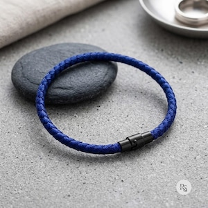 Pode incluir: Uma pulseira de cordão trançado em azul royal com um fecho cilíndrico de metal preto. A pulseira tem formato circular e apresenta uma textura tecida consistente em todo o cordão, fixada por um mecanismo de fecho liso e de tom escuro.