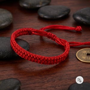 May include: A bright red woven cord bracelet featuring a braided pattern and adjustable sliding knots. The bracelet is constructed from a single continuous strand of cord, creating a circular band with two loose ends extending from the knot mechanism.