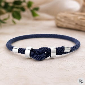 May include: A navy blue rope bracelet with silver metal details. The bracelet has a knot in the centre and is secured with two silver metal rings. A stylish accessory.