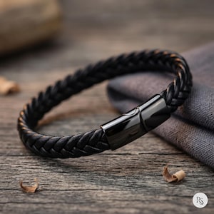 May include: A black braided leather bracelet with a black magnetic clasp. The bracelet is displayed on a wooden surface with a gray cloth in the background. The bracelet's design features a woven pattern.