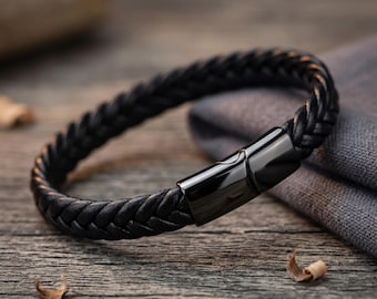 Könnte beinhalten: Ein schwarzes geflochtenes Lederarmband mit einem schwarzen Magnetverschluss. Das Armband wird auf einer Holzoberfläche mit einem grauen Tuch im Hintergrund präsentiert. Das Design des Armbands weist ein geflochtenes Muster auf.