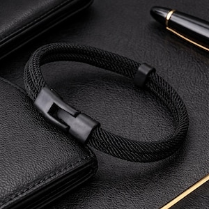 Peut inclure: Bracelet en corde tressée noire avec fermoir noir mat. Le bracelet est posé sur une surface en cuir noir à côté d'un portefeuille noir et d'un stylo aux accents dorés. Un accessoire élégant.