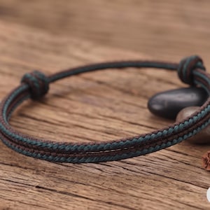 Könnte beinhalten: Ein zweisträngiges Kordelarmband mit einem geflochtenen Muster in Dunkelbraun und Blaugrün. Das Armband ist mit zwei verstellbaren Schiebeknoten auf beiden Seiten konstruiert, was eine flexible Anpassung an das Handgelenk ermöglicht.