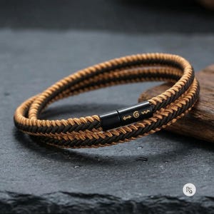 Puede incluir: Una pulsera de doble vuelta con dos cordones trenzados entrelazados en color tostado y marrón oscuro. La pulsera se asegura con un cierre cilíndrico de metal negro que muestra una pequeña flecha, una espiral y una línea ondulada doradas.