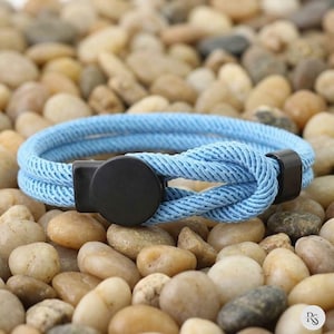 Può includere: Un braccialetto di corda azzurra con chiusura nera. Il braccialetto è realizzato in un materiale intrecciato e strutturato ed è posto su uno sfondo di piccole pietre lisce. Un accessorio semplice ma elegante.
