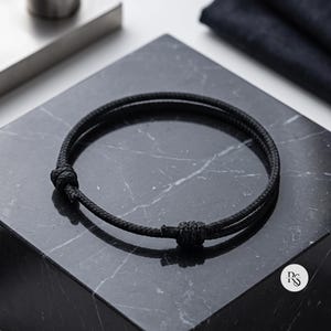 May include: A black cord bracelet featuring a double-strand design with two adjustable sliding knots. The bracelet is constructed from a textured, woven material and maintains a circular shape, showcasing a simple and functional aesthetic.