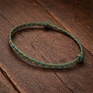 Peut inclure: Un bracelet en corde vert et or avec une fermeture nouée. Le bracelet est fait d'une matière de corde tissée avec un motif répété de vert et d'or. Le bracelet est présenté sur une surface en bois.