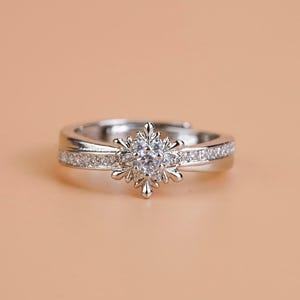 Puede incluir: Un anillo de tono plateado con un adorno central en forma de copo de nieve engastado con una piedra transparente facetada. La banda está pulida e incluye una hilera de pequeñas piedras transparentes brillantes incrustadas en los laterales.