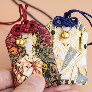 May include: Two rectangular fabric omamori amulets with hanging cords and small gold bells. One is deep red with black and gold architectural patterns, while the other is white with blue and gold accents. Both feature intricate woven designs.