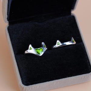 Könnte beinhalten: Zwei silberne Ringe mit Katzenohren-Design, jeweils mit einem grünen herzförmigen Edelstein. Die Ringe werden in einer grauen Samt-Ringbox mit schwarzem Innenraum vor einem pfirsichfarbenen Hintergrund präsentiert.