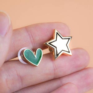 Puede incluir: Dos pequeños pines de metal, uno con forma de corazón con relleno de esmalte verde y el otro con forma de estrella de cinco puntas con acabado metálico dorado, que incluyen un cierre de plástico transparente en el pin de corazón.