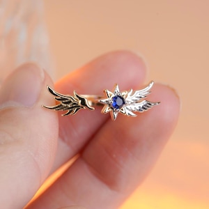 Puede incluir: Un pendiente de oreja dorado y plateado con diseño de ala. Un extremo tiene un ala pequeña y el otro una estrella con una gema azul en el centro y alas. La joya se sostiene entre los dedos.