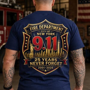 Może przedstawiać: Granatowa koszulka z krótkim rękawem z grafiką na plecach. Wzór zawiera tarczę ze złotą obwódką z napisem 'FIRE DEPARTMENT CITY OF NEW YORK 9.11', '25 YEARS NEVER FORGET' i '2001-2026' oraz wizerunkiem Statuy Wolności.