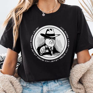 Könnte beinhalten: Schwarzes T-Shirt mit einem kreisförmigen Schwarz-Weiß-Motiv eines Mannes mit Hut, der mit dem Finger zeigt. Der Schriftzug "JETZT REDE ICH" umrundet das Motiv. Das T-Shirt ist aus weichem Material.