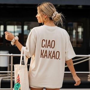 Könnte beinhalten: Cremefarbenes, übergroßes T-Shirt mit dem Aufdruck "CIAO KAKAO" in Braun auf dem Rücken. Die Person hält eine Tragetasche mit einem bunten Design. Die Person trägt ein passendes Scrunchie und goldene Creolen.