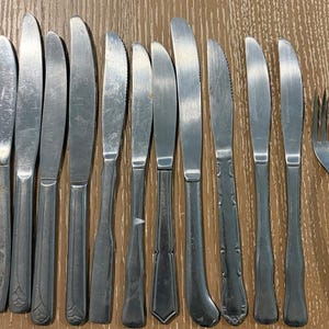 Puede incluir: Una colección de cuchillos de acero inoxidable y un tenedor están dispuestos sobre una superficie de madera. Los cuchillos varían en diseño, algunos con bordes dentados y mangos decorativos. El tenedor está a la derecha.