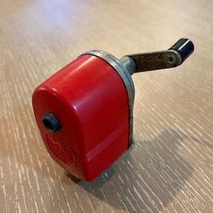 Pode incluir: Um apontador de lápis vermelho vintage com uma faixa de metal prateado e uma pega preta. O apontador tem uma forma retangular com bordas arredondadas e um pequeno orifício para o lápis. A pega está presa na lateral.