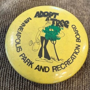 Pode incluir: Botão amarelo com o texto "ADOPT A TREE" e "MINNEAPOLIS PARK AND RECREATION BOARD". O botão apresenta um desenho animado de uma pessoa regando uma árvore com um rosto sorridente. O botão é redondo.